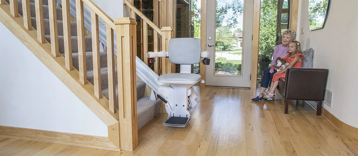 Bruno Elite straight indoor stairlift parked at the bottom of a staircase next to a grandmother and little girl