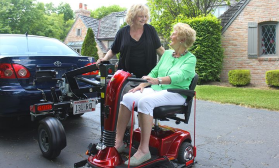 an elderly women parking a scooter onto a scooter lift mounted on the back of a blue car