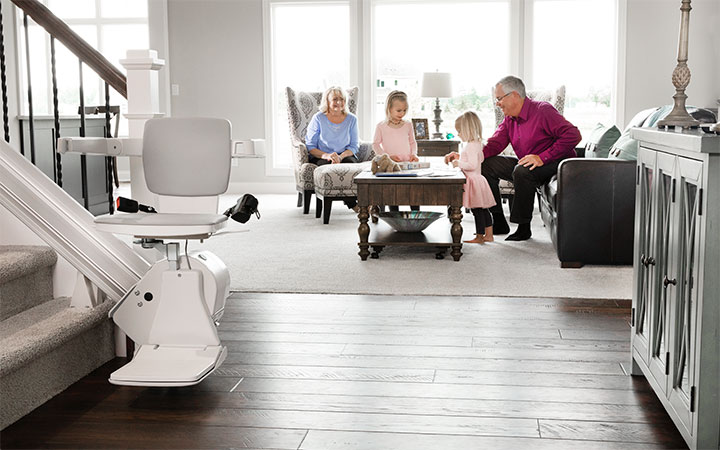 Woman riding a Bruno indoor stair lift at the bottom of a staircase with two girls smiling on the steps