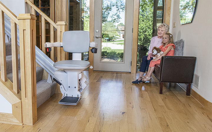 Bruno Elite straight indoor stairlift parked at the bottom of a staircase next to a grandmother and little girl