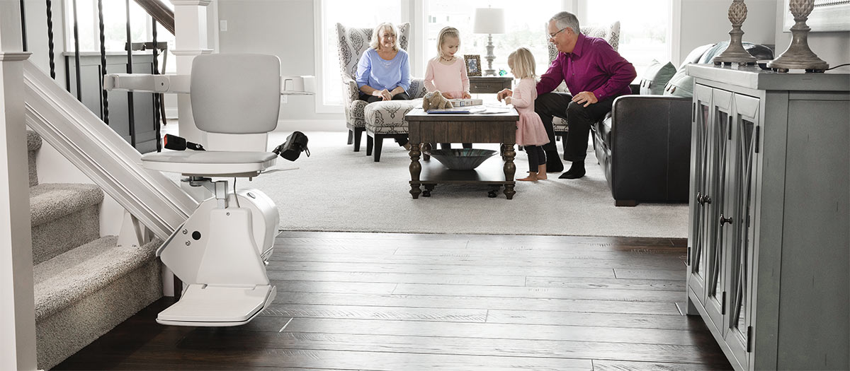 Woman riding a Bruno indoor stair lift at the bottom of a staircase with two girls smiling on the steps